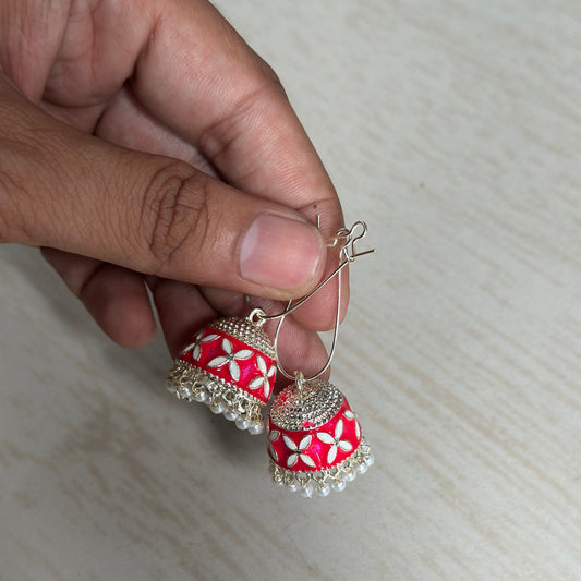 Red and silver jhumka-style earrings adorned with white floral patterns and pearl hangings.